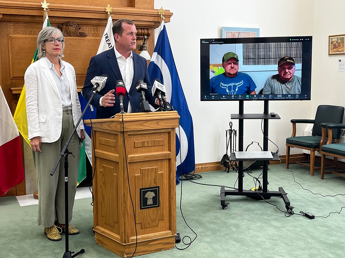 Deputy agriculture critic Trent Wotherspoon and municipal affairs critic Erika Ritchie stand at a lectern during a news conference. Two men in ballcaps are visible on a small screen, joining the conference remotely, to the speakers' left.