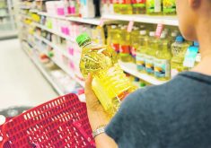 A shopper holds a clear plastic container of golden vegetable oil in her hand and looks at it in the aisle of a grocery store.