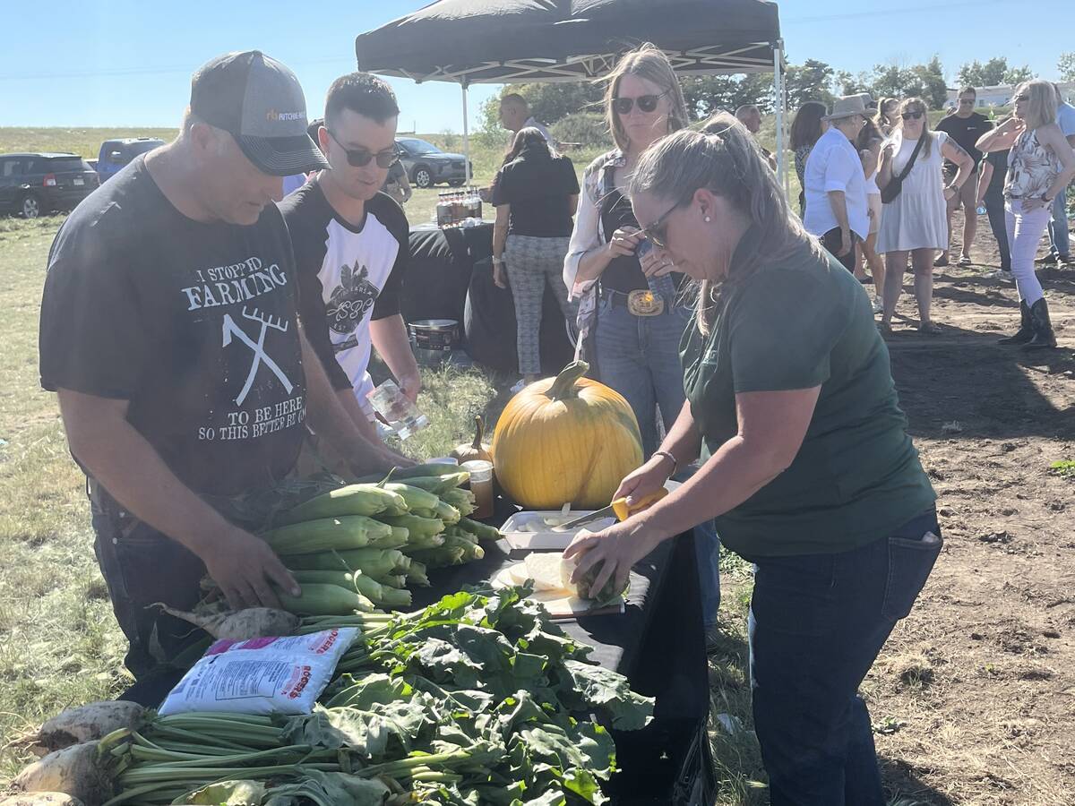 A man and a woman stand over a table loaded with fresh produce, including corn and a pumpkin.