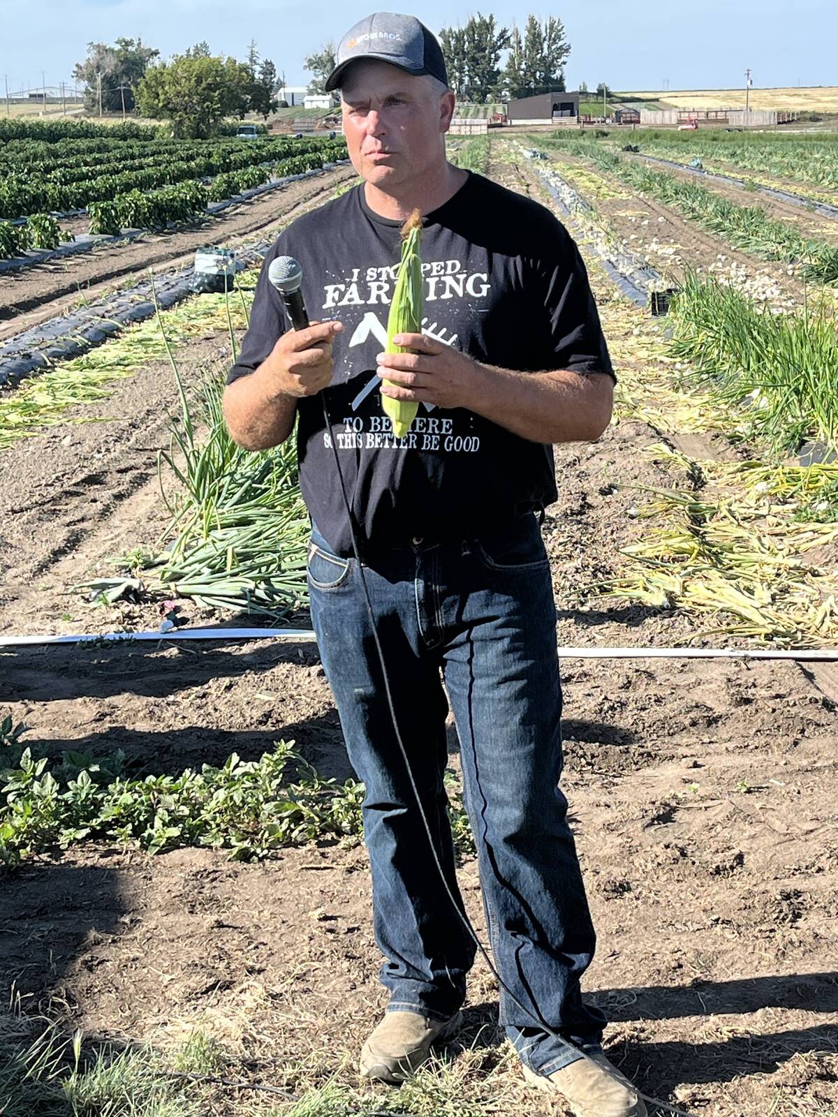 A man in a ball cap, black t-shirt and blue jeans stands in front of a large vegetable garden holding a microphone in one hand and an ear of corn in the other.