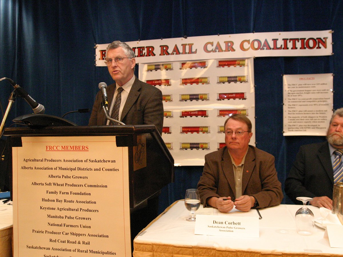 An archive photo of Sinclair Harrison speaking at a podium with a "Farmer Railcar Coalition" sign on the wall behind him.