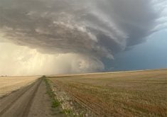 A storm front featuring dark clouds is clearly visible over prairie fields with a gravel road running toward the storm along the left side of the image.