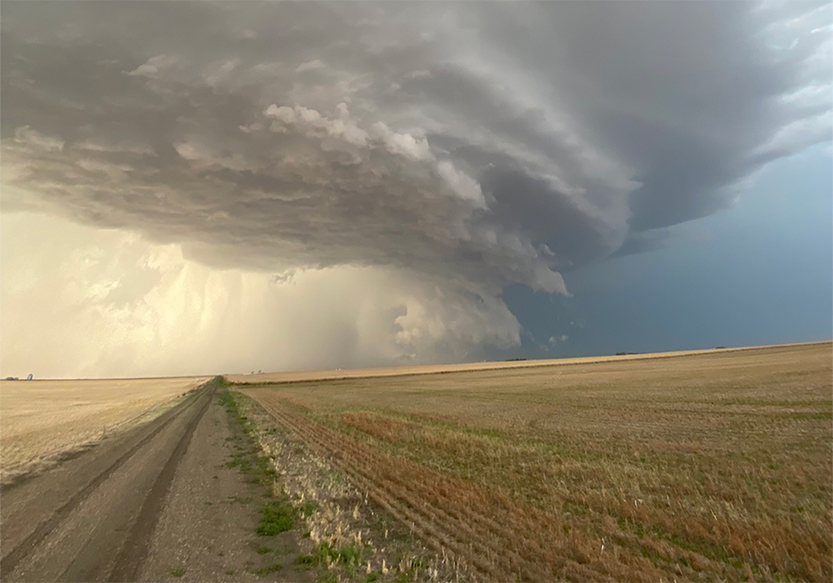 A storm front featuring dark clouds is clearly visible over prairie fields with a gravel road running toward the storm along the left side of the image.