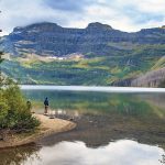 Two people stand on the shore of Cameron Lake, in Waterton Lakes National Park, looking across the water at the mountains in the distance.