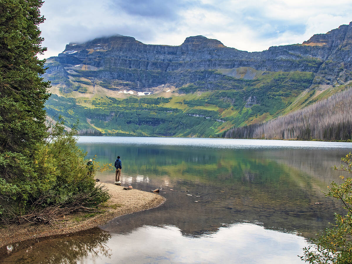 Two people stand on the shore of Cameron Lake, in Waterton Lakes National Park, looking across the water at the mountains in the distance.