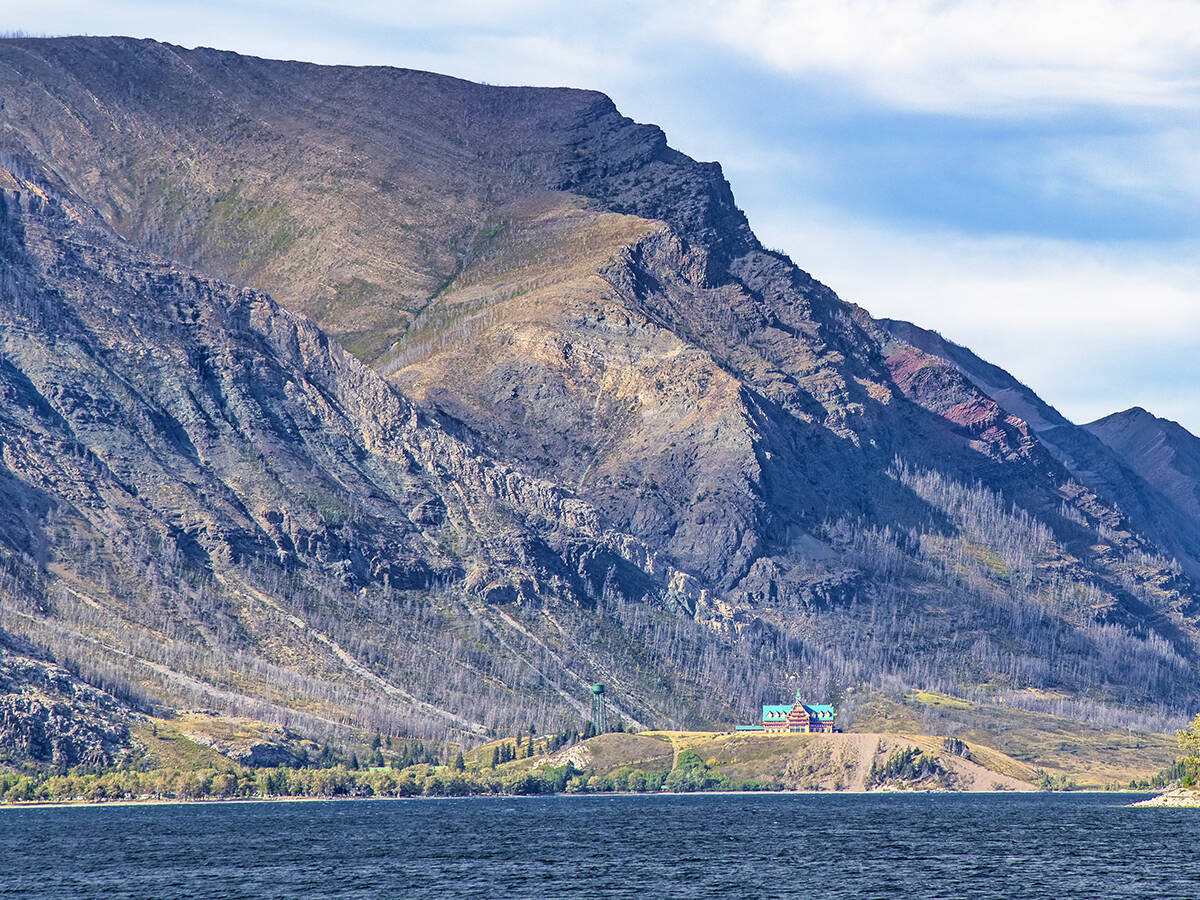 The Prince of Wales Hotel on Upper Waterton Lake is seen from a distance across the lake with mountains rising in the background.