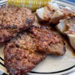 Two pieces of ranch steak, a cob or corn and some quartered potatoes, with skin still on and some pan gravy poured overtop, sit on a plate.