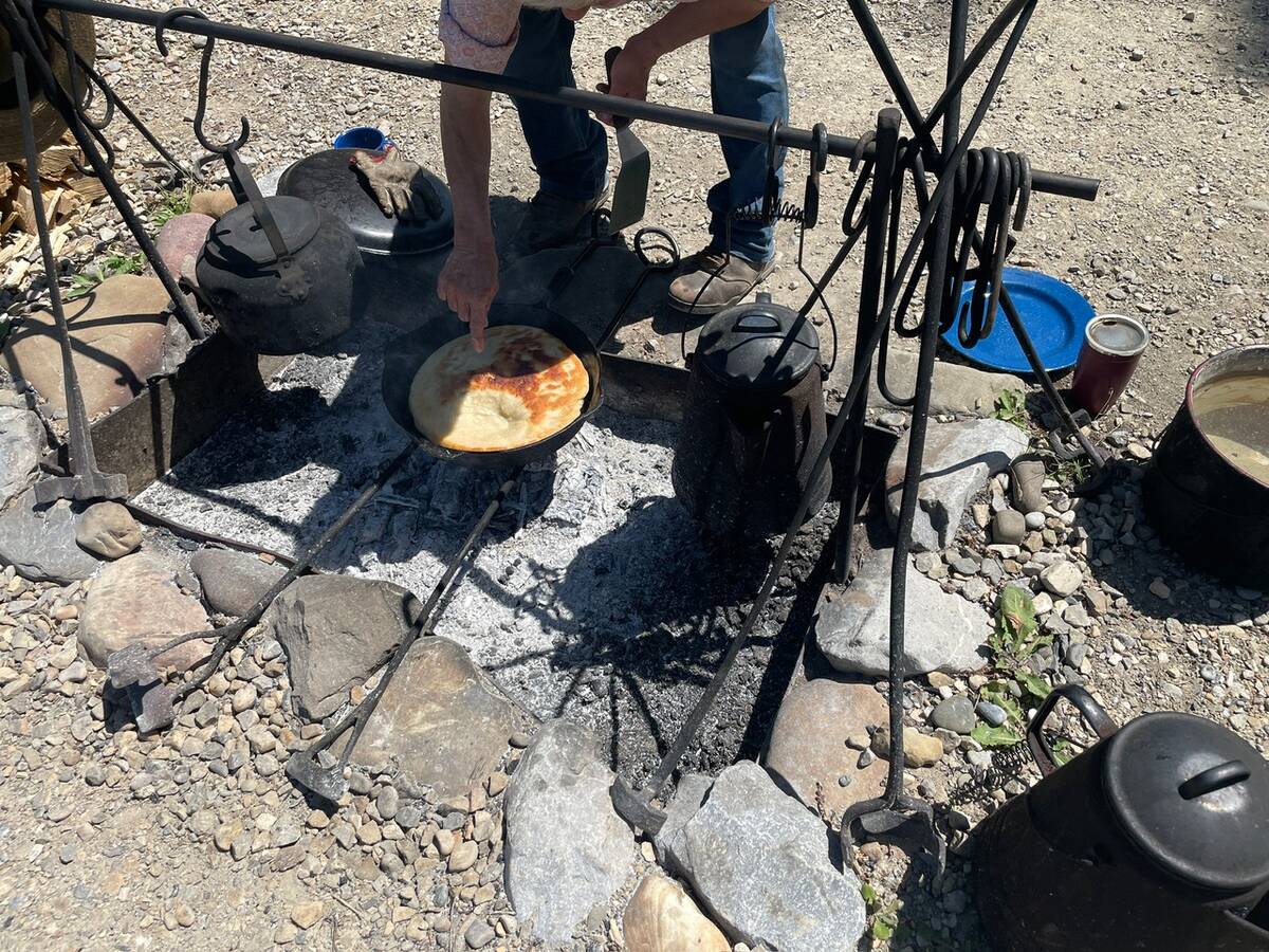 A man leans over a cast iron frying pan checking on the bannock roasting over wood coals in an outdoor camp-style fire pit.
