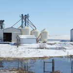 A seed cleaning plant is surrounded by melting snow and above a slough in spring time.