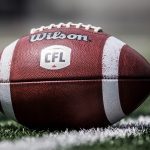Close-up of a Wilson CFL leather football sitting on the green turf with a white painted stripe beneath the football on the turf.