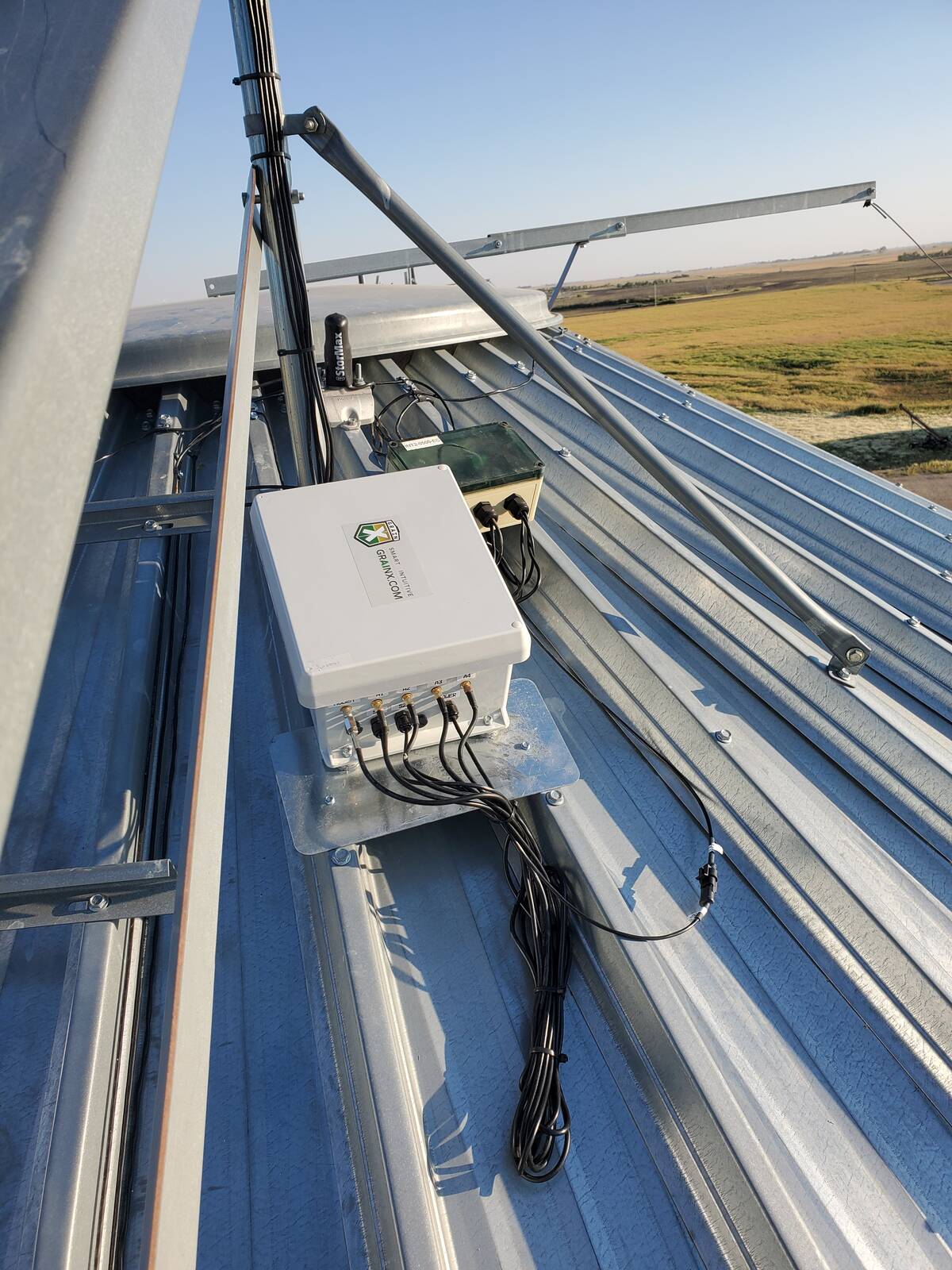 A GrainX AI unit, a gray metal box with about 8 black wires coming out of it, is mounted on a bin roof.