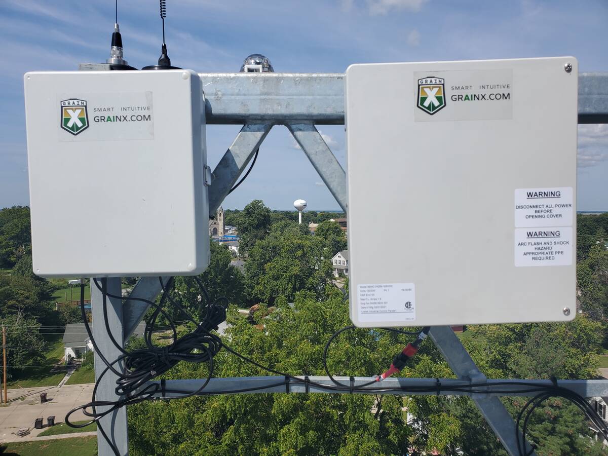 Two gray metal boxes, the power supply and AI unit are attached to a railing on a grain bin high above the ground.