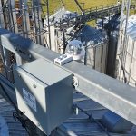 A GrainX rain sensor and power supply box is shown high up on a grain bin with other smaller bins in the background.