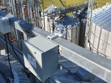 A GrainX rain sensor and power supply box is shown high up on a grain bin with other smaller bins in the background.