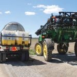 A sprayer is parked on the side of a gravel road being loaded by a tanker truck carrying a pesticide tank mix.