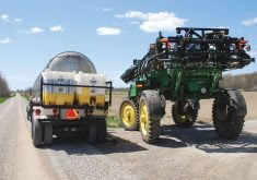 A sprayer is parked on the side of a gravel road being loaded by a tanker truck carrying a pesticide tank mix.
