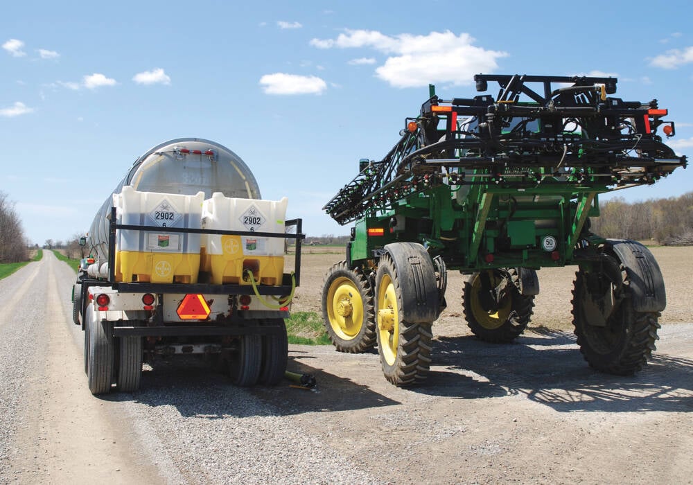A sprayer is parked on the side of a gravel road being loaded by a tanker truck carrying a pesticide tank mix.