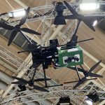 Looking upward at the bottom of a large John Deere spray drone with two propellers on each of its four arms, that is suspended over the floor of a trade show display booth.