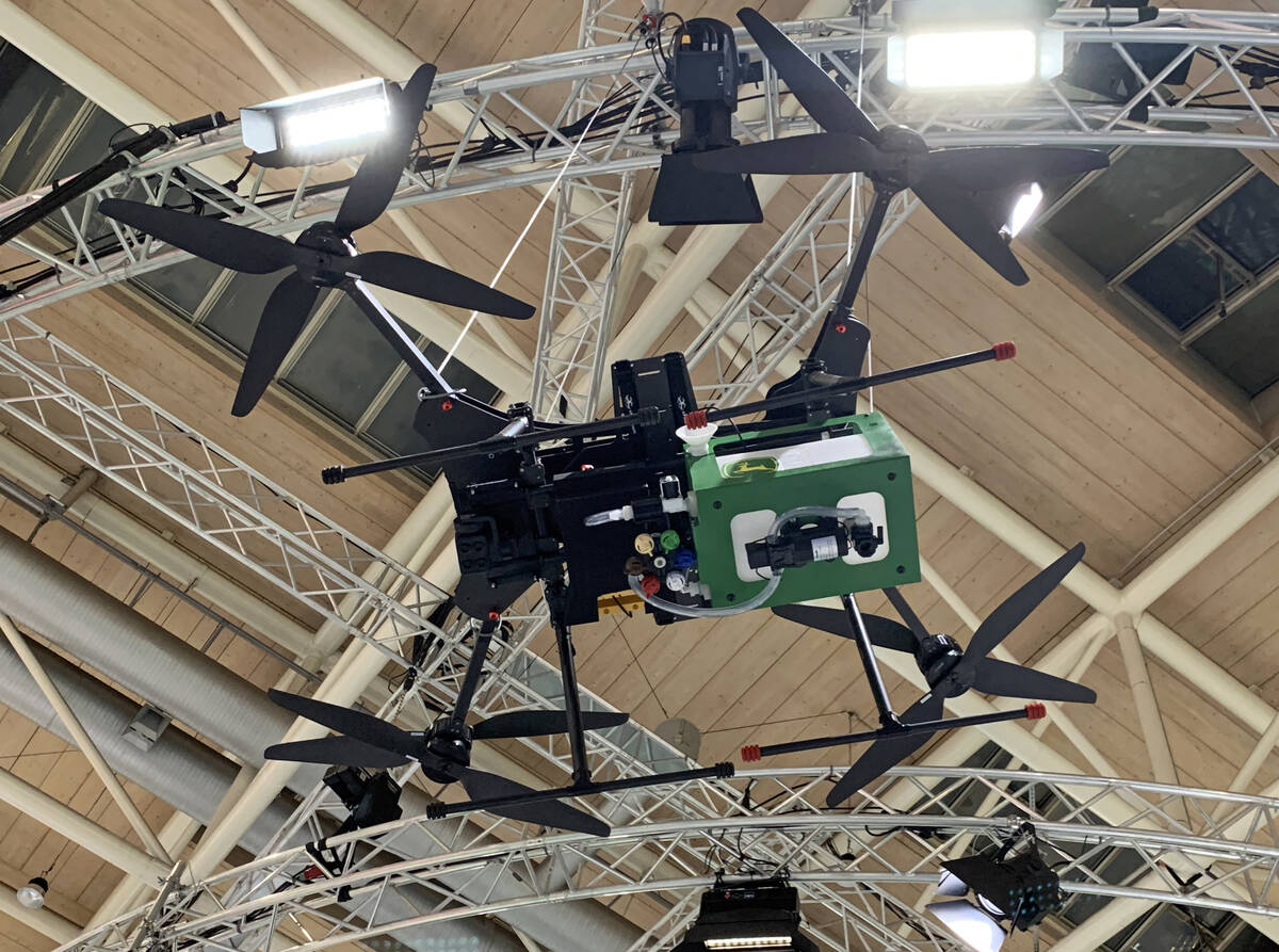 Looking upward at the bottom of a large John Deere spray drone with two propellers on each of its four arms, that is suspended over the floor of a trade show display booth.