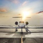 A stock image looking straight at the nose of a private jet sitting on a runway with the sun setting behind it.