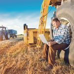 A man sits on the hub of a tire on a large piece of farm equipment with a laptop open and resting on his knees.