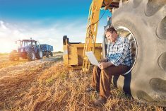 A man sits on the hub of a tire on a large piece of farm equipment with a laptop open and resting on his knees.