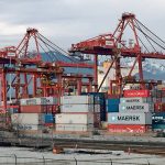 Stacks of shipping containers sit dockside beneath the massive cranes that hoist them aboard ships in the Port of Vancouver with the mountainous North Shore visible in the background.