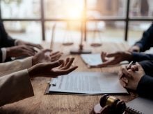 The animated hands of people seated on opposite sides of a boardroom table with a gavel in the foreground, and a blurry "scales of justice" statue in the background.