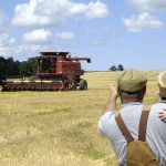 An older male farmer holds a young, blonde child and points toward a red combine in a recently-harvested wheat field about 40 metres away.