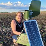 A woman kneels in a potato field next to a round, green metal cylinder - a spore trap - mounted on a tripod that has a solar panel on it.