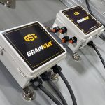 Close-up of a two gray boxes mounted on the sloped roof of a metal grain bin with "GSI GrainVue" written on them, each with multiple black cables entering and exiting.