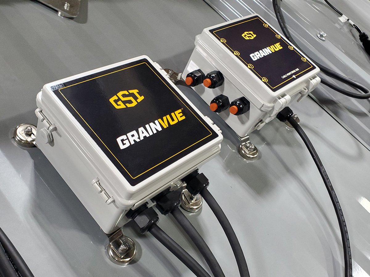 Close-up of a two gray boxes mounted on the sloped roof of a metal grain bin with