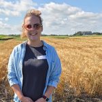A woman in a light blue denim shirt with a dark blue shirt underneath poses for a photo, smiling, in front of a wheat field.