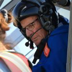 An older man sits in the pilot seat of a helicopter wearing a helmet with a radio mike boom attached.