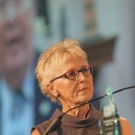 Dori Gingera-Beauchemin stands near a podium and microphone with a large screen behind her.