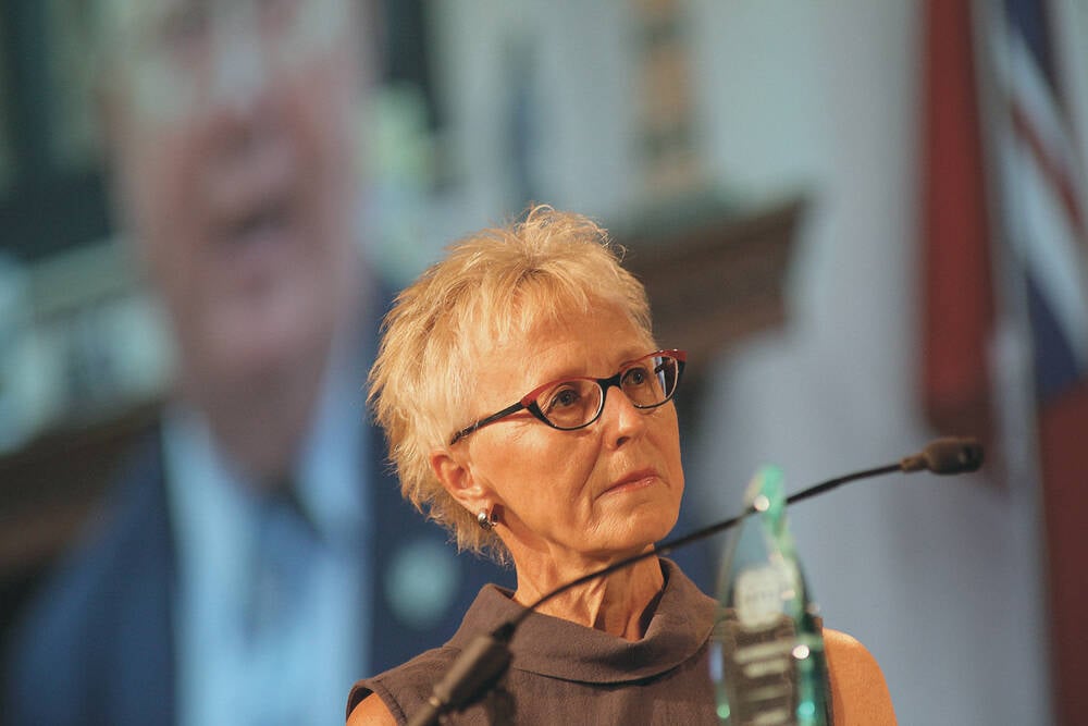 Dori Gingera-Beauchemin stands near a podium and microphone with a large screen behind her.