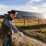 A man in a black cowboy hat wearing work gloves and a vest with a tool belt over his blue jeans stands in front of a large solar array.