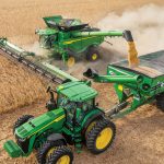 A stock photo provided by John Deere showing, from above, a combine harvesting corn and augering it into a grain cart being pulled by a tractor beside it.