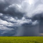 A stock photo of some dark storm clouds with an obvious downpour happening over farm land in the middle of the image.