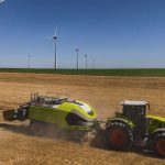 A promotional drone photo of the Class 70 t sqare baler in action in a field beneath a number of large wind turbines.