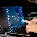 A man's hands are on the keyboard of a laptop and its screen is folded up beyond vertical and a hologram is being projected between the laptop's screen and keyboard which one of the hands on the keyboard is poking to select something with its index finger.