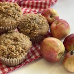 Several freshly-baked muffins sit next to a few apples on a red and white checkered tablecloth.