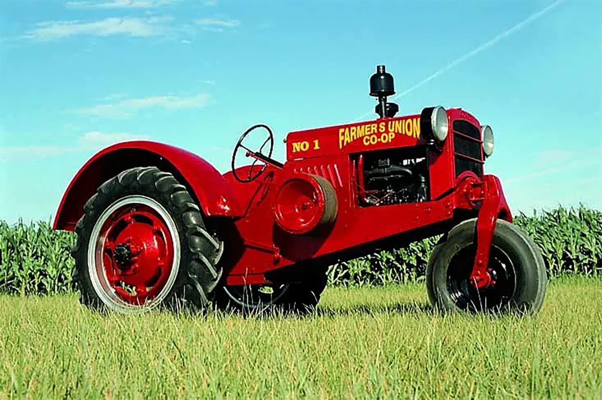 A low angle photo of a red "Co-Op" tractor parked on the gras with a corn crop behind it on a sunny summer day with a blue sky.