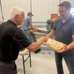 One man holds a "frame" from a bee hive with fresh honeycombs and honey on it for him to sample inside a white tin-walled processing facility.