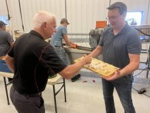 One man holds a "frame" from a bee hive with fresh honeycombs and honey on it for him to sample inside a white tin-walled processing facility.