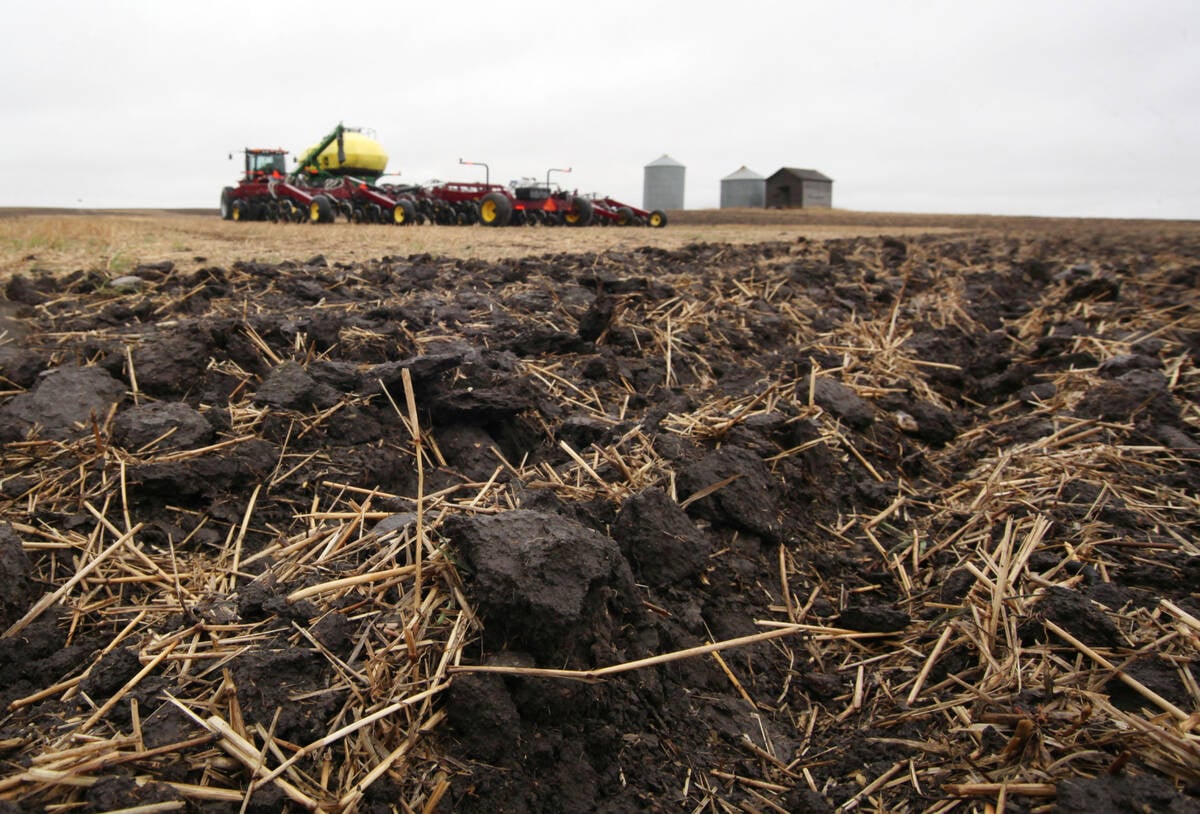 A strip of worked soil in a Prairie field with a seeder and fertilizer farm equipment set up in the background during seeding season.