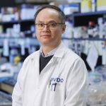 A scientist in a white lab coat with "VIDO" (an acronym for the Vaccine and Infectious Disease Organization) poses for a photo in a lab with shelves packed with lab-related materials and a crowded countertop blurred in the background behind him.