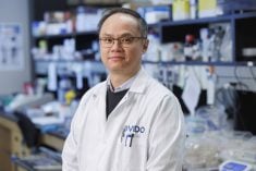 A scientist in a white lab coat with "VIDO" (an acronym for the Vaccine and Infectious Disease Organization) poses for a photo in a lab with shelves packed with lab-related materials and a crowded countertop blurred in the background behind him.