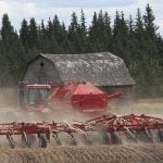 A tractor kicks up dust as it pulls a seeder planting winter wheat with an old barn and some pine trees in the background.