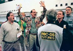 A trader on the floor of the Winnipeg Commodity Exchange stands, clad in a jacket with a busy pattern, hands raised above his head with a pen in one hand and a receipt-like book in the other, as other traders stand around him.